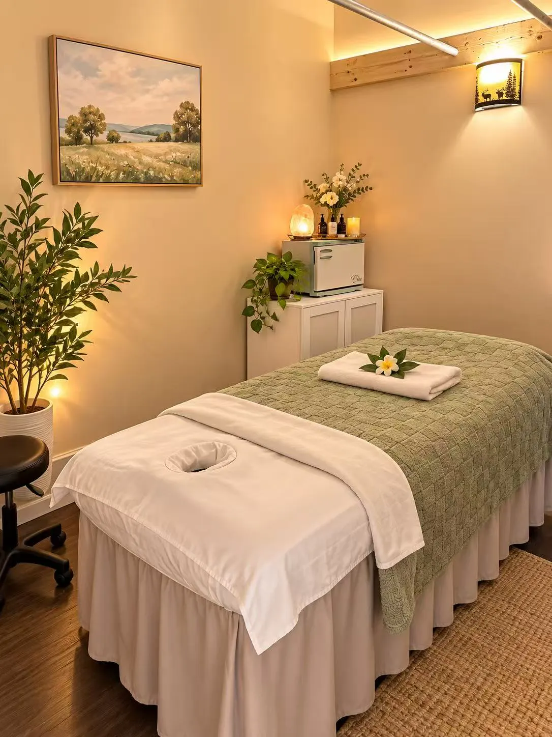Warmly lit treatment room: a massage table with a sage blanket, a wall sconce and a jute rug