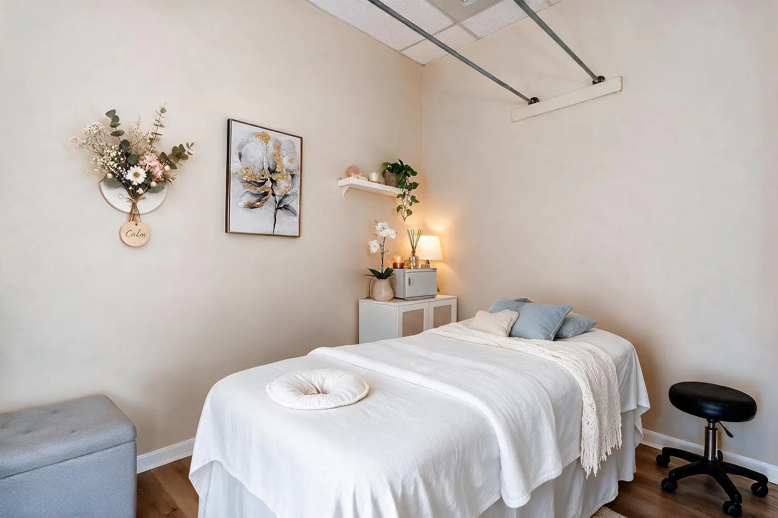 A cream-walled treatment room with a massage table in white linens, blue pillows and a lamp